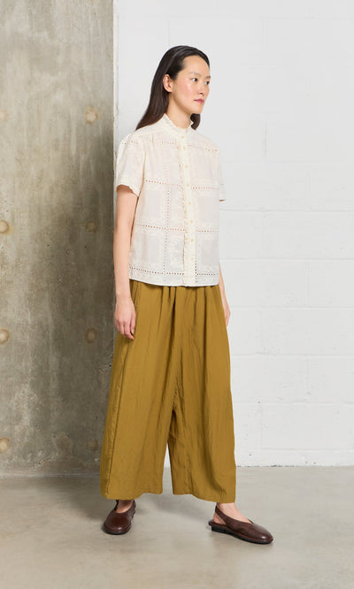 Plümo- Woman stands indoors in a cream blouse, Carrick wide-leg mustard pants with pleated waistband, and dark brown slip-ons.