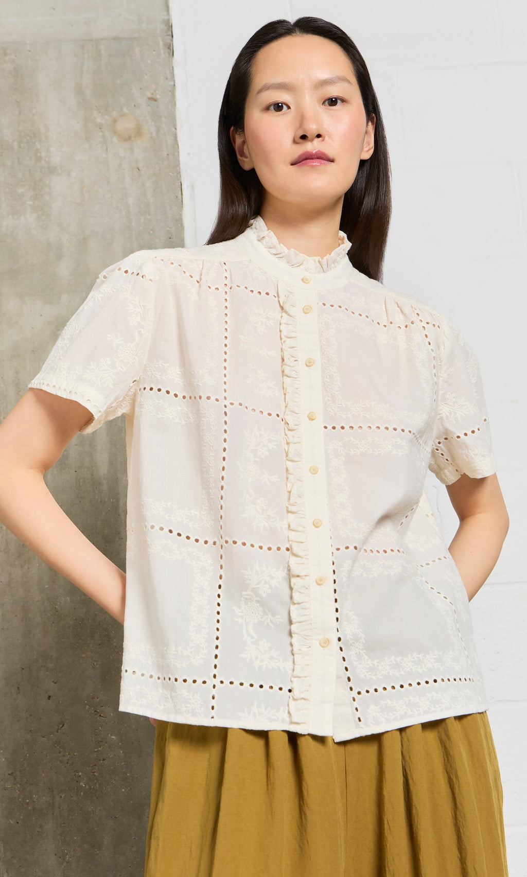 Plümo- Woman wears the Gill Blouse, a short-sleeve broderie anglaise top with ruffle trim, by a concrete and white wall.