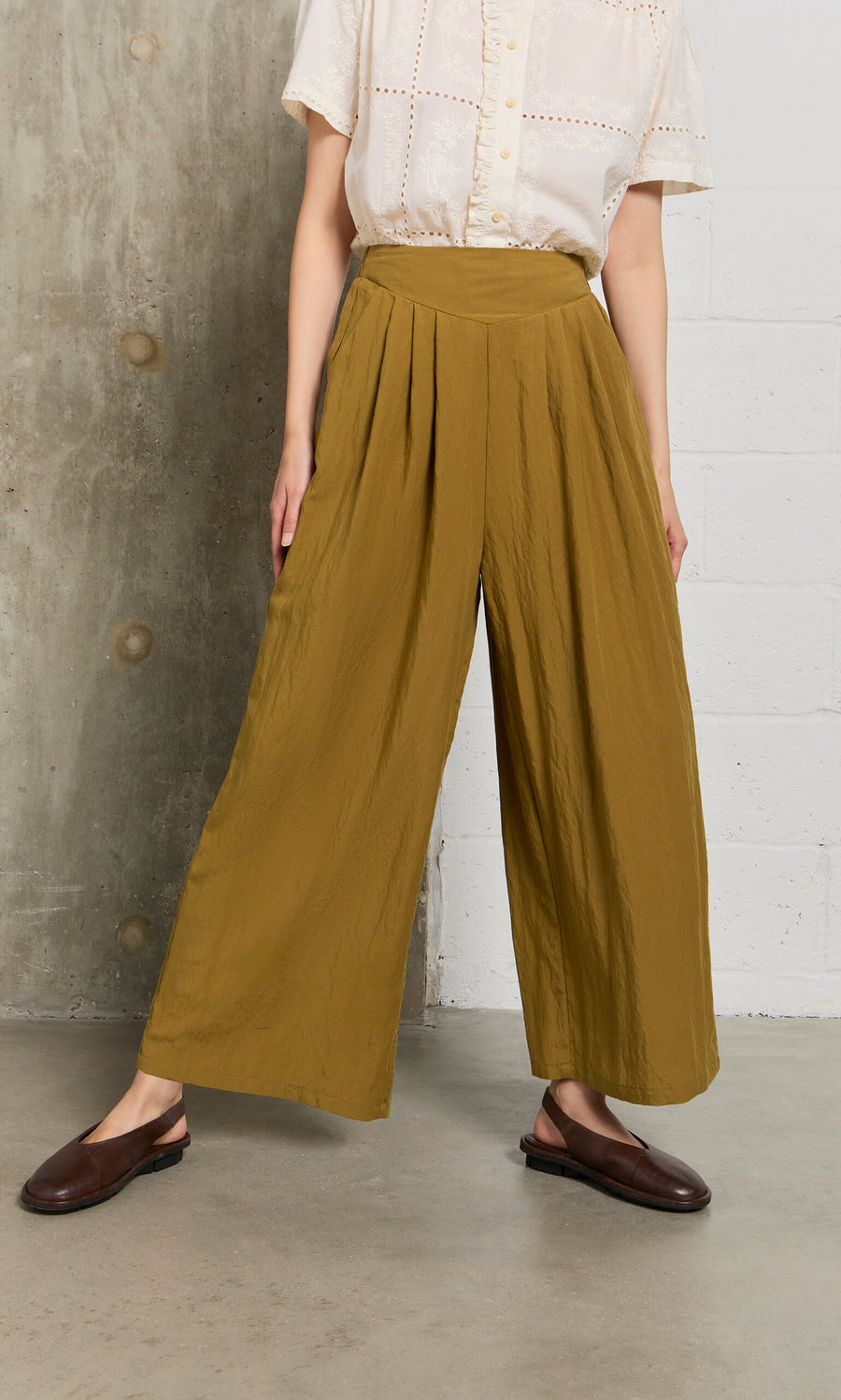 Plümo- Person wears Carrick Pants with a pleated waistband, a cream blouse, and brown shoes against a textured wall.