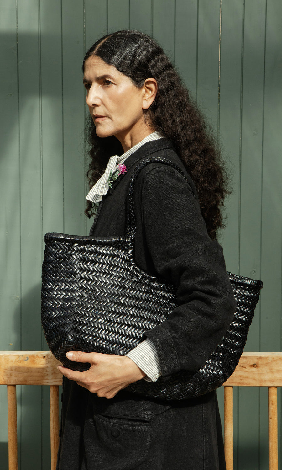 Plümo  Woman in a black coat holds the Lyle Bag, a basket-weave leather tote with braided handles, by a green wooden wall.