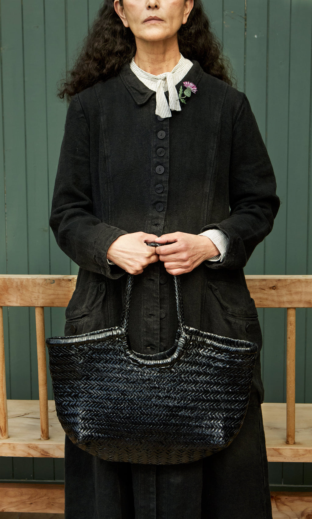 Plümo  Woman in a black dress with the spacious Lyle Bag, posing by a green wooden wall.