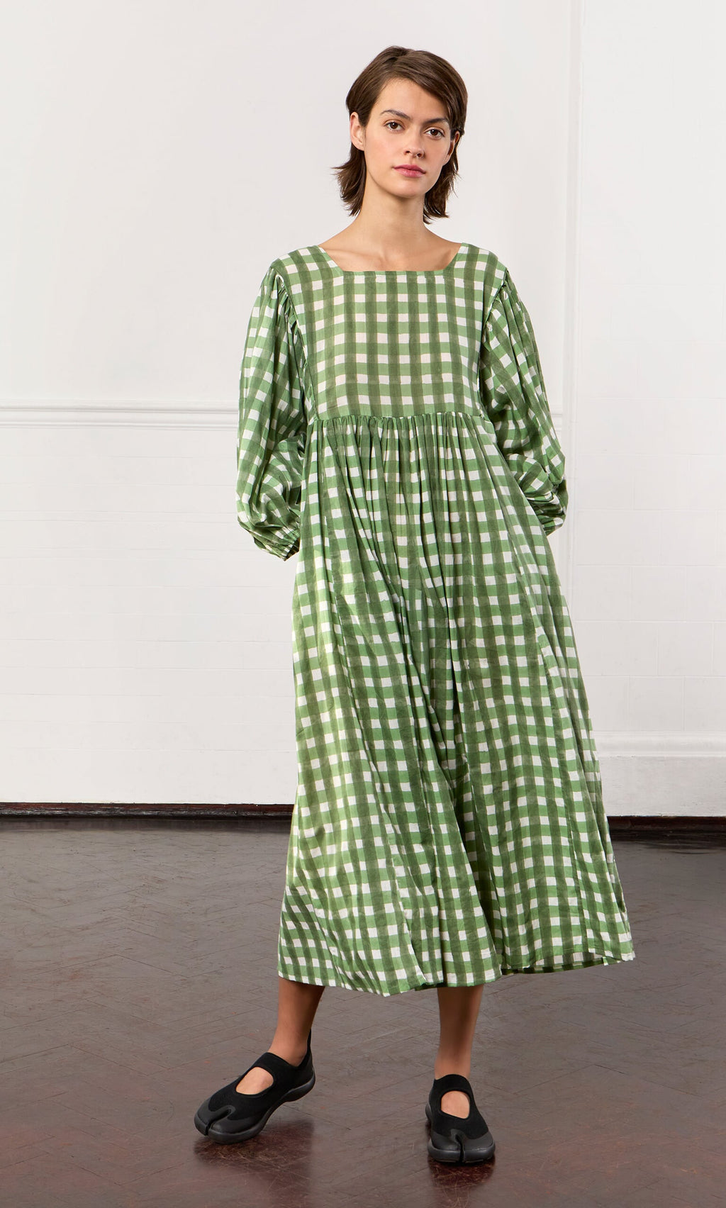 Plümo- Woman wearing the Marta Dress, a hand-printed gingham folk cotton dress, stands indoors in black shoes.
