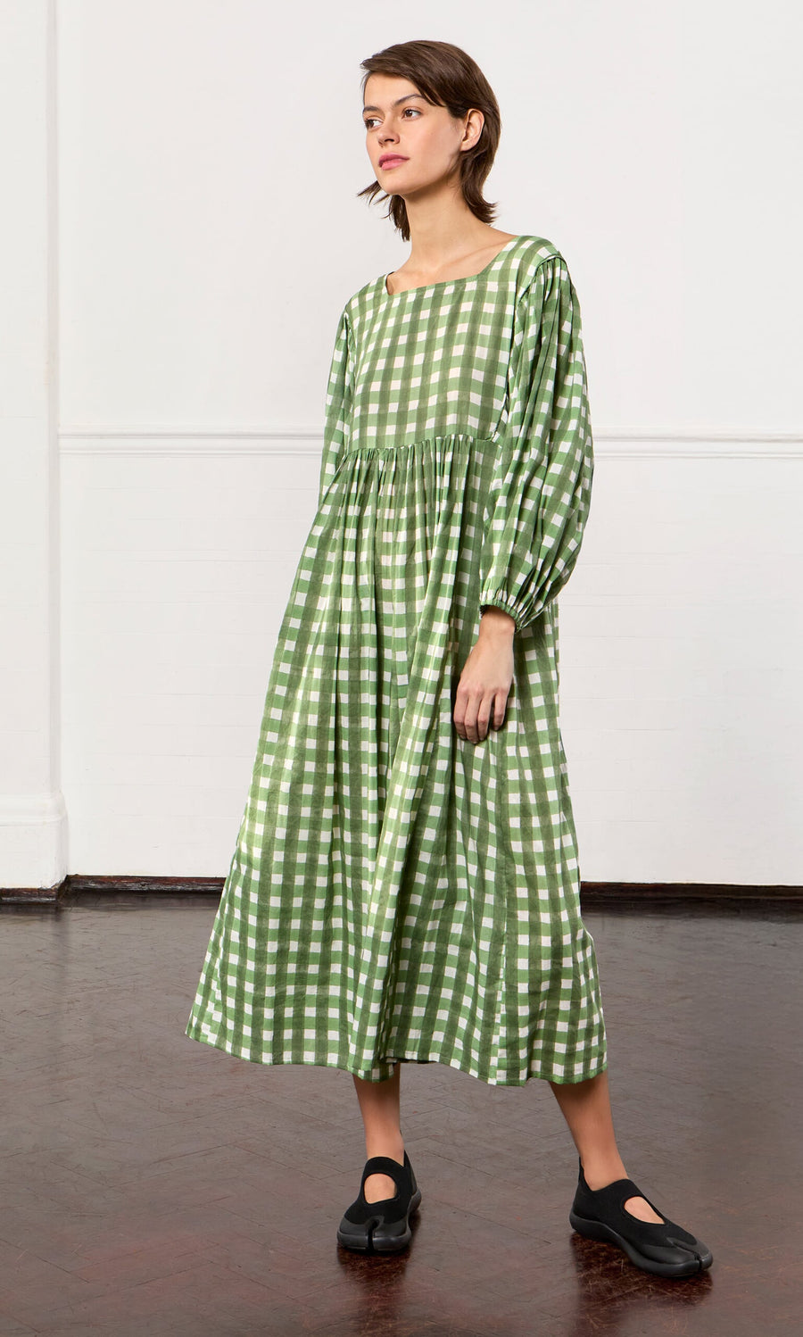 Plümo- Person in a Marta Dress—a long, hand-printed gingham folk cotton dress—wears black shoes indoors on a dark floor.