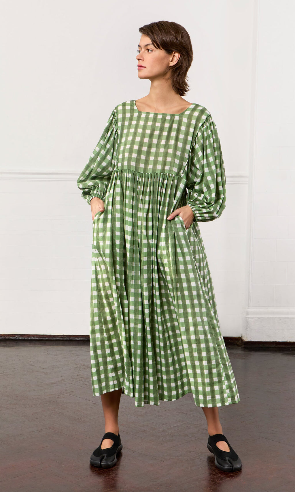 Plümo- Woman wearing the Marta Dress, a hand-printed cotton gingham dress with pockets, standing on a dark wood floor.
