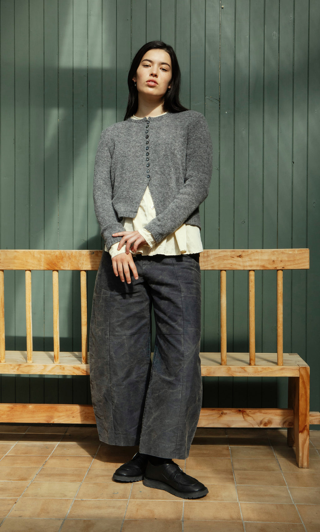 Plümo  Woman wearing the Mawgan Cardigan stands in sunlight beside a wooden bench and a green paneled wall.