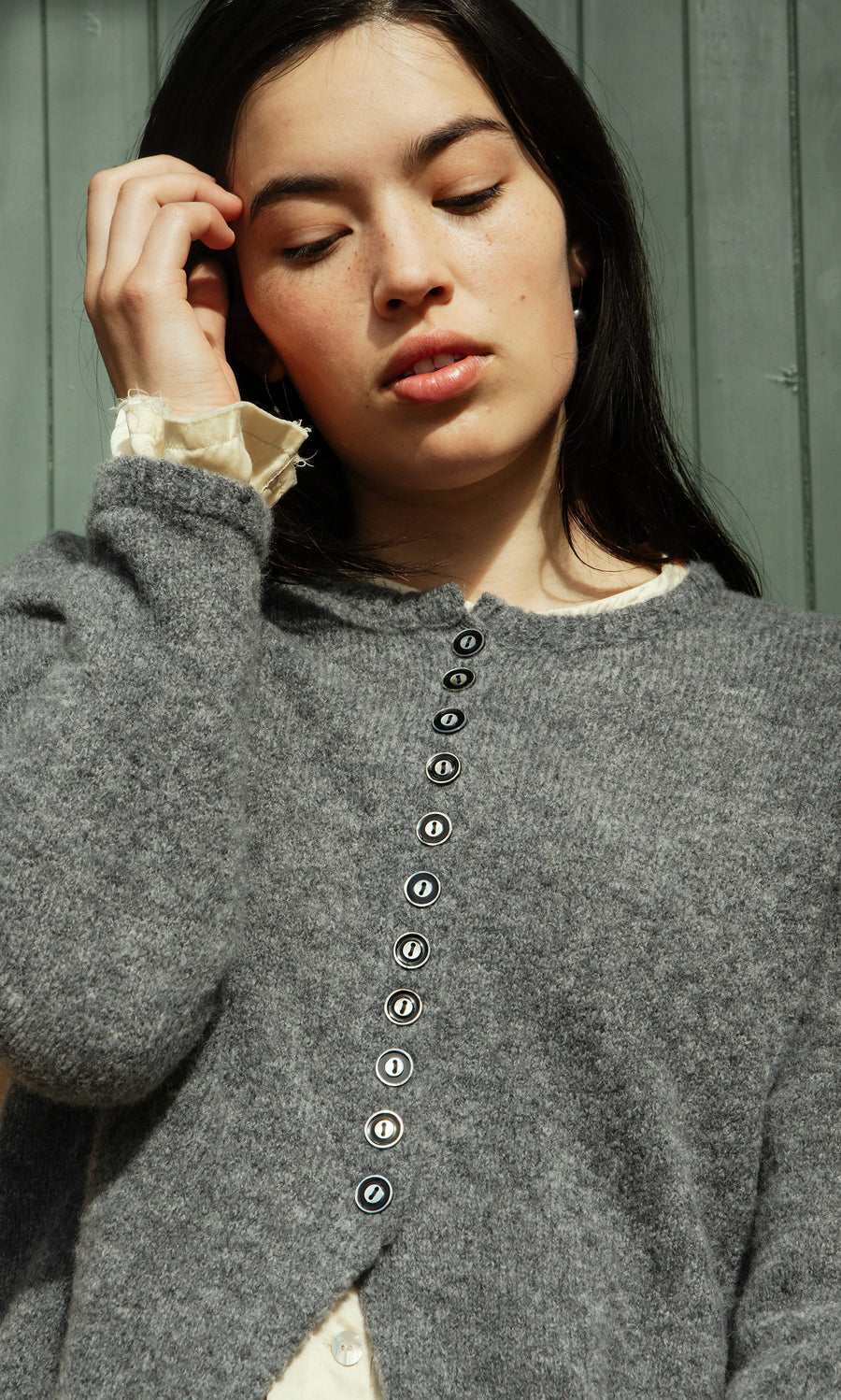 Plümo  Woman with long dark hair wears the Mawgan Cardigan with enamelled shell buttons, near green wooden panels.
