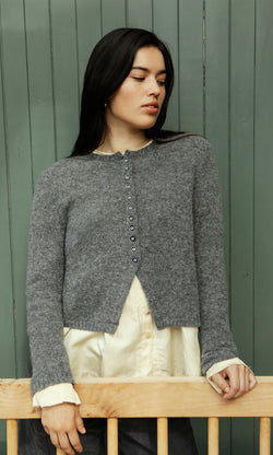 Plümo  Woman in the Mawgan Cardigan stands by a green wooden wall, gazing thoughtfully to the side.