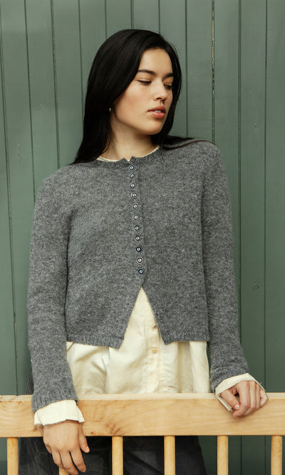 Plümo  Woman in the Mawgan Cardigan stands by a green wooden wall, gazing thoughtfully to the side.