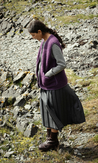 Plümo  A woman in a Maylin Gilet and gray skirt stands on rocky terrain, hands in her vest pockets, looking down.