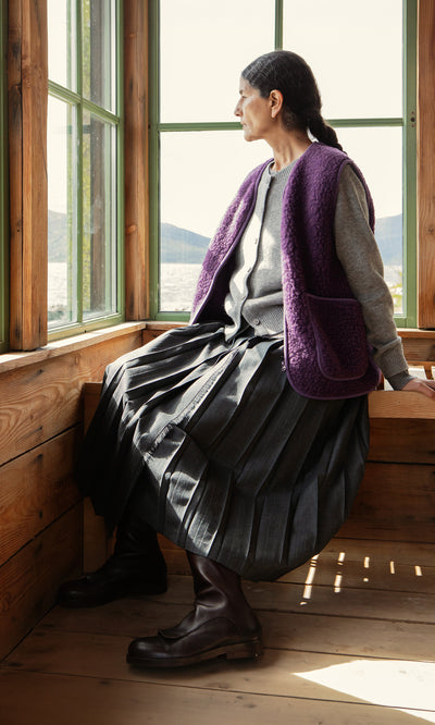 Plümo  Woman in pleated skirt and Maylin Gilet sits on a window seat, gazing out at the lake and mountains.