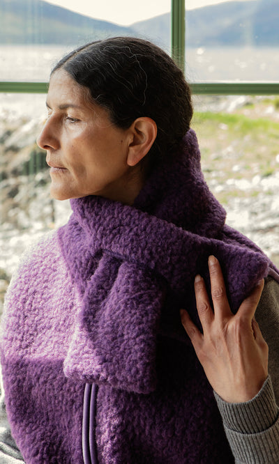 Plümo  Woman in a thick purple scarf, gray sweater, and Maylin Gilet, gazing out a scenic window.