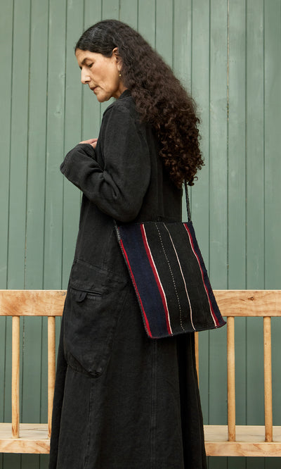 Plümo  Woman in a long black coat with the Medina Bag stands by a wooden bench in front of green panels.