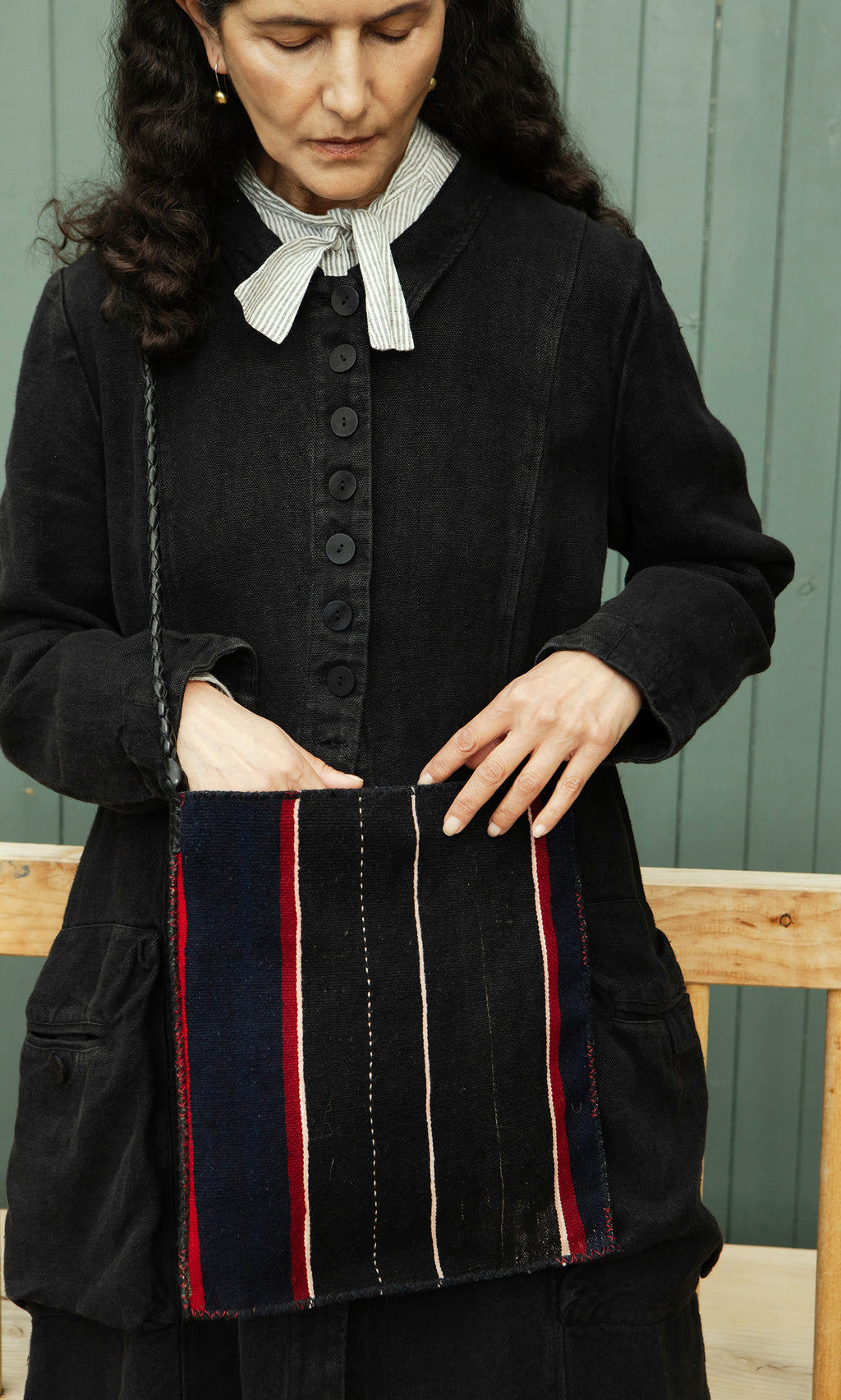 Plümo  Woman wears the Fowey Coat, relaxed-fit black linen, holding a striped bag in front of a green wall.