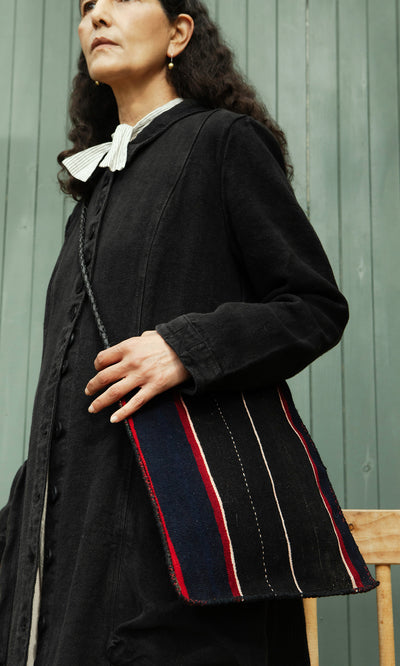 Plümo  Woman in a black coat holds the Medina Bag with red, blue, and white lines, standing by a green wall.