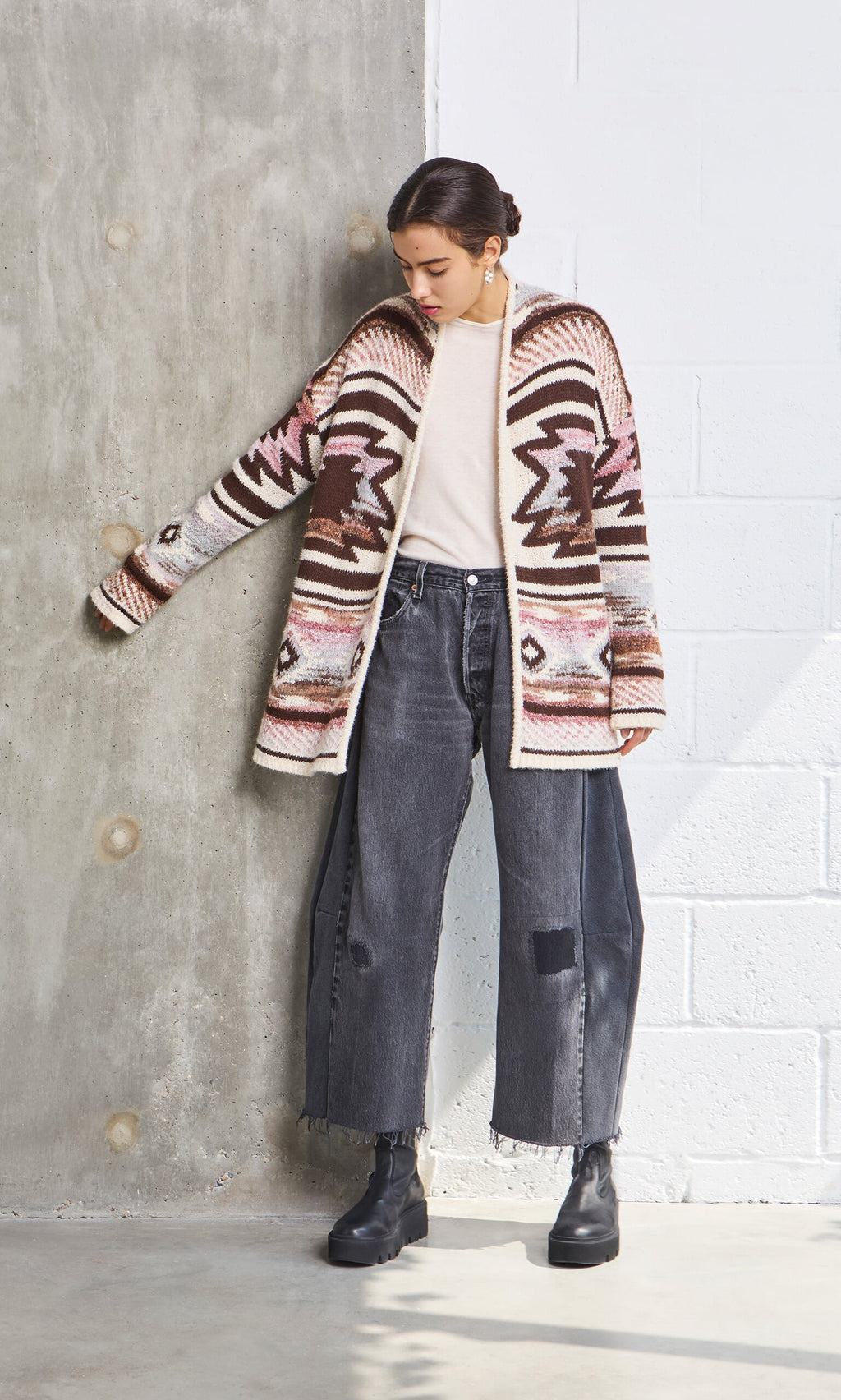 Plümo- Woman in a Mika Cardigan, light top, black jeans with patch, and boots stands by a concrete wall.