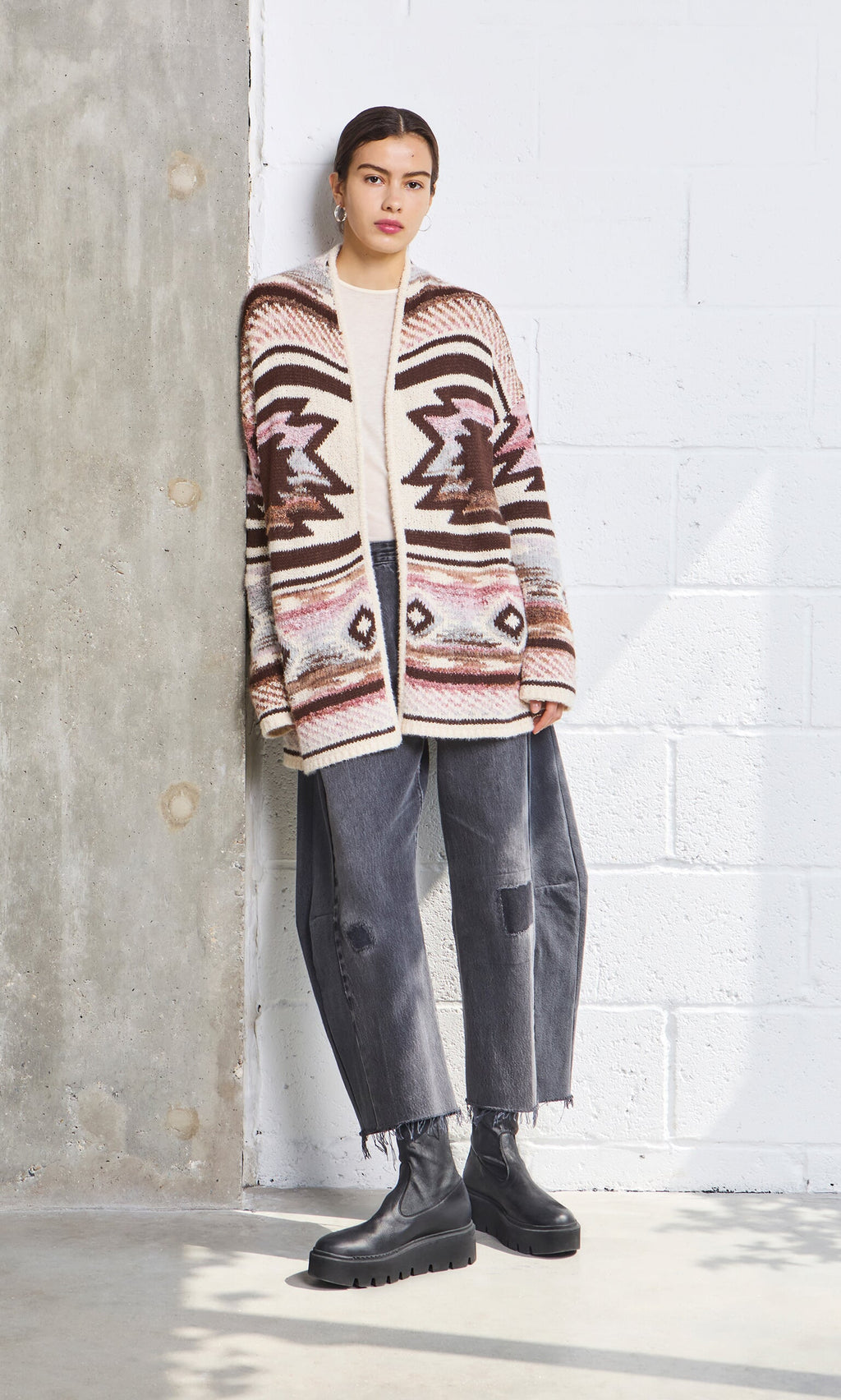 Plümo- Woman standing by a concrete wall, wearing the Mika Cardigan, black jeans, and black boots.
