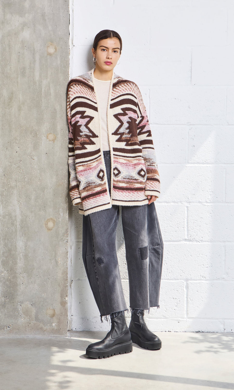 Plümo- Woman standing by a concrete wall, wearing the Mika Cardigan, black jeans, and black boots.