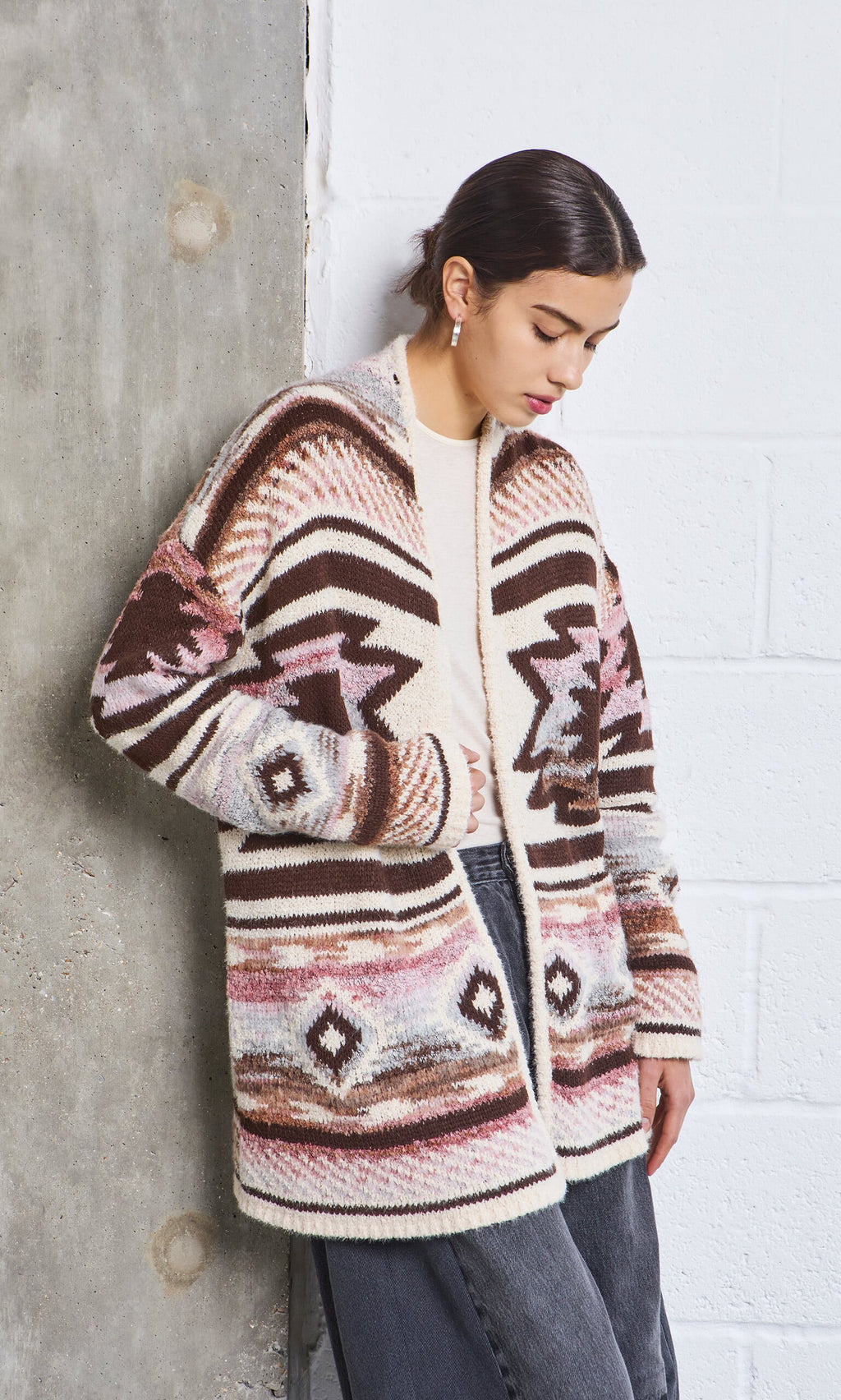 Plümo- Woman in the Mika Cardigan and black jeans stands against a concrete and white brick wall, looking down.