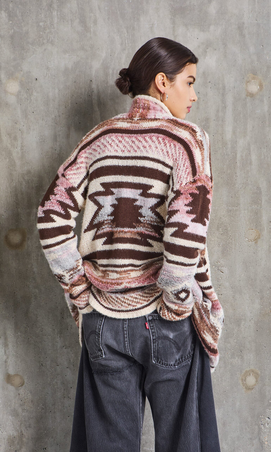 Plümo- Woman with dark hair in a bun wears the Mika Cardigan and black jeans, standing against a concrete wall, facing away.