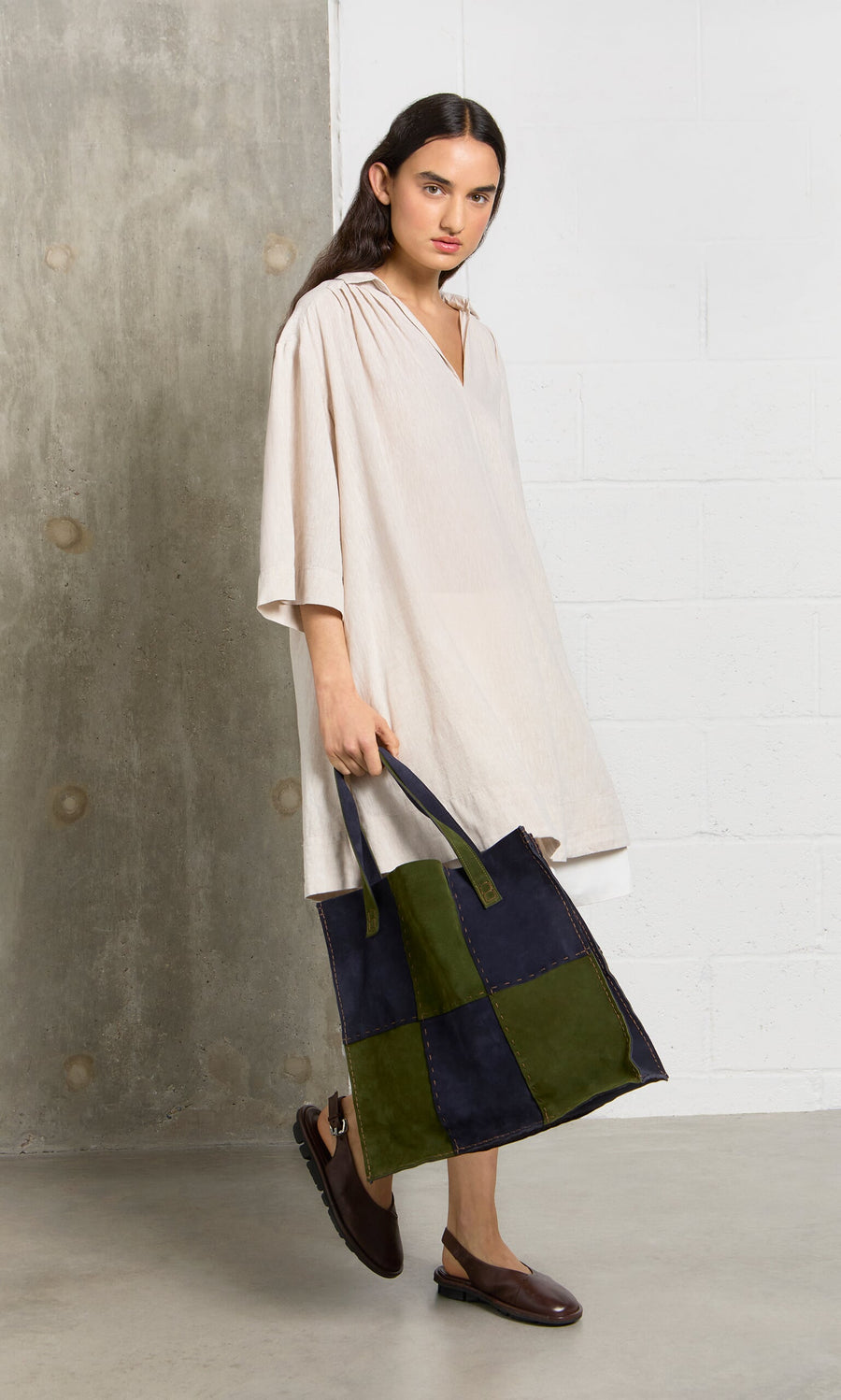 Plümo- Person in a loose beige dress holding the Patchwork Suede Bag, standing indoors on a concrete floor.