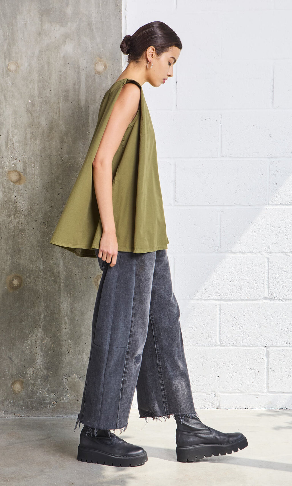 Plümo- Woman in a Mina Top - Calke Green, wide-leg gray jeans, and chunky boots walks indoors.
