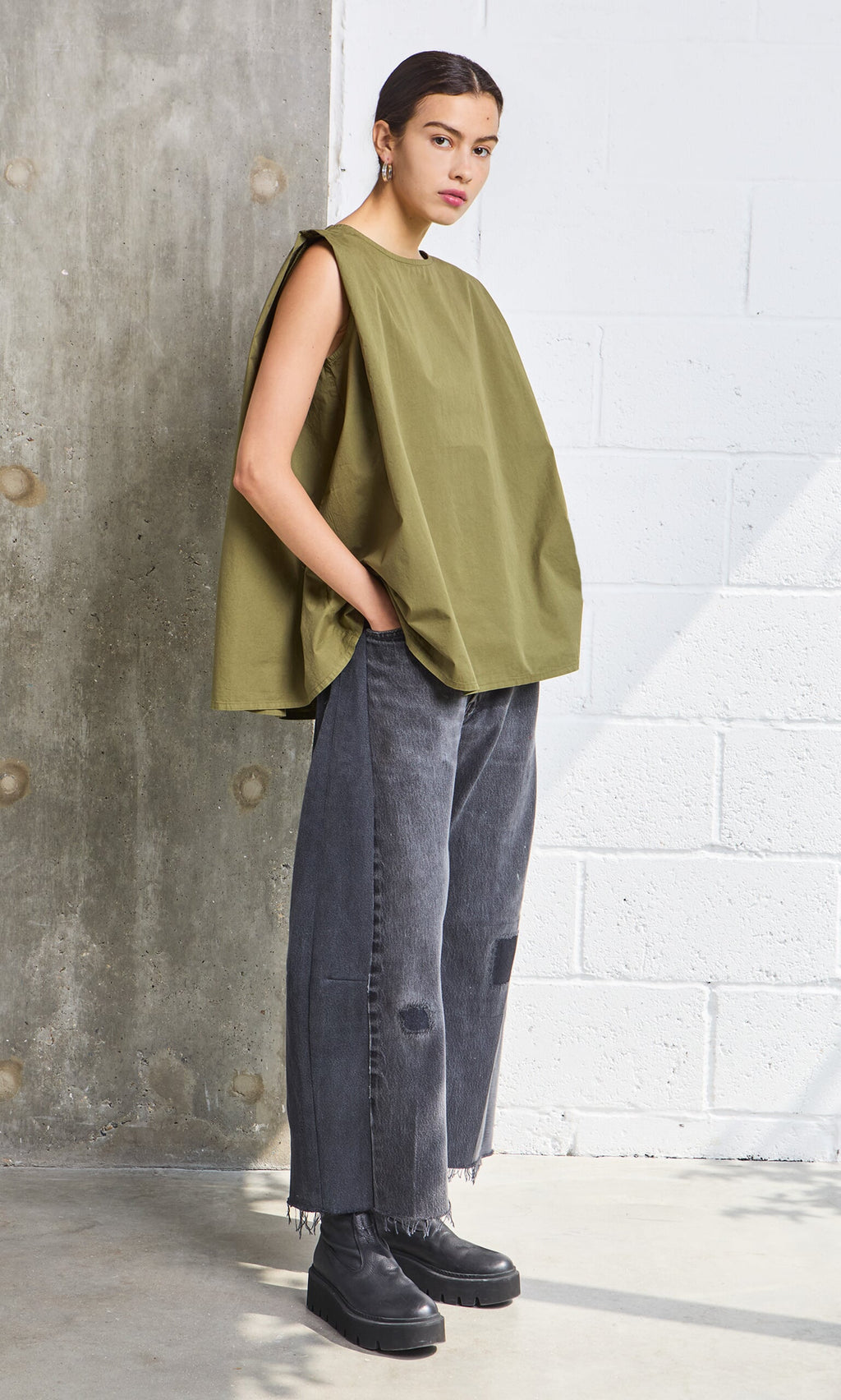 Plümo- Woman in Mina Top - Calke Green, wide gray jeans, and black boots stands against a brick wall.