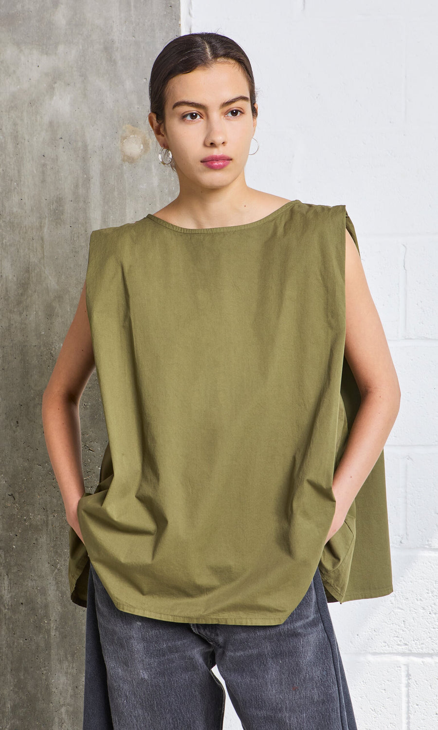 Plümo- Woman in the Mina Top - Calke Green and gray jeans stands against a concrete and white wall—PLÜMO Studio style.