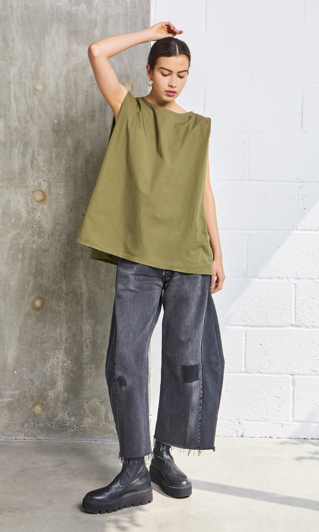 Plümo- A person in the Mina Top - Calke Green and wide-leg gray jeans stands against a concrete and white brick wall.