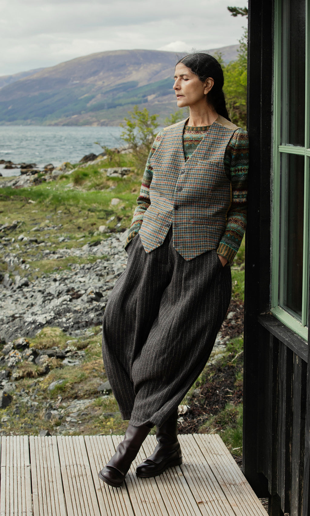 Plümo  A person in a Moorland Waistcoat stands on a porch by a lake, gazing thoughtfully into the distance.