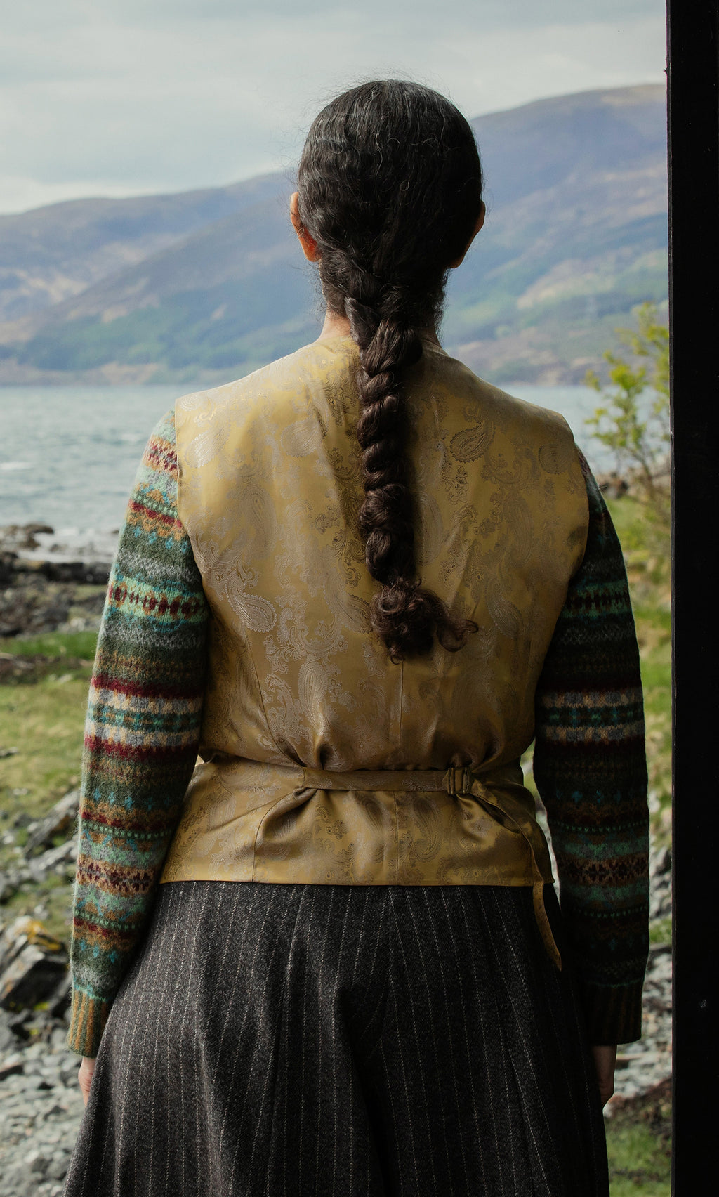 Plümo  A person with a long braid faces a lake and mountains, wearing the Moorland Waistcoat and a patterned sweater.