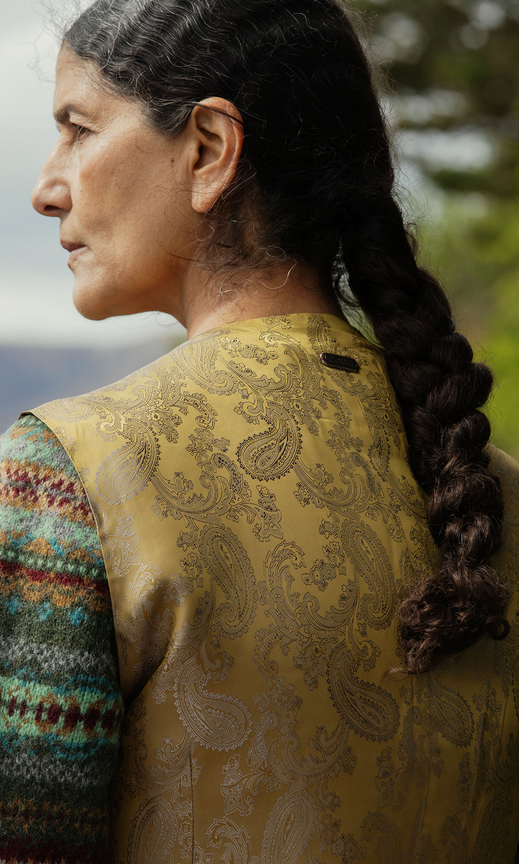 Plümo  A woman with a long braid wears the Moorland Waistcoat in gold paisley, standing outdoors, back to the camera.