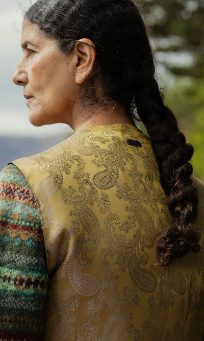 Plümo  A woman with a long braid wears the Moorland Waistcoat in gold paisley, standing outdoors, back to the camera.
