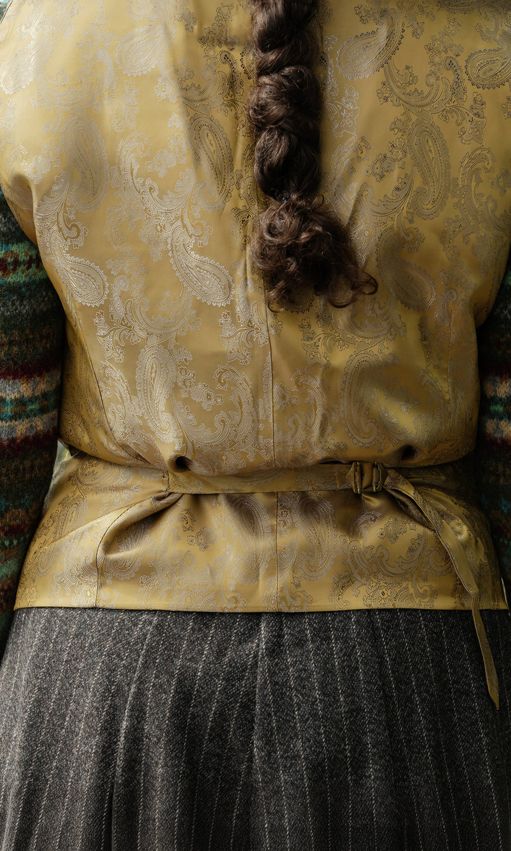 Plümo  A person with a braided ponytail wears a Moorland Waistcoat and striped skirt, seen from behind.