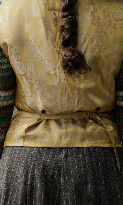 Plümo  A person with a braided ponytail wears a Moorland Waistcoat and striped skirt, seen from behind.