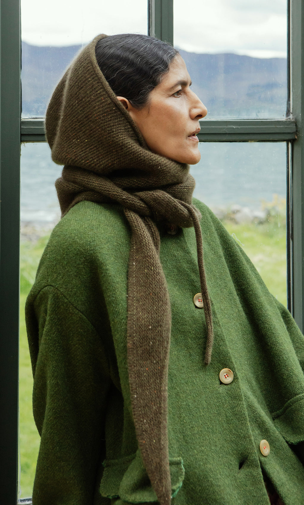 Plümo  Woman in green coat and Moss Specked Hood stands by window, gazing at the mountains.