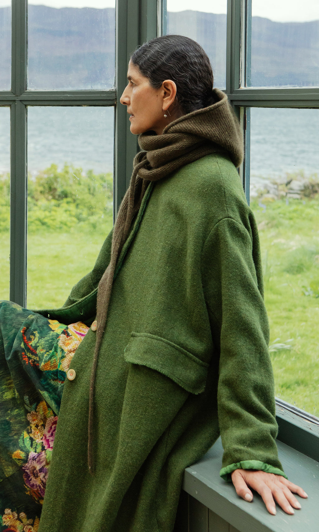 Plümo  Woman in a Moss Specked Hood sits by a window, gazing at water and faraway green mountains.