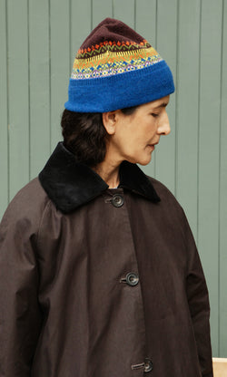 Plümo  Person in a Mull Fairisle Beanie and dark coat stands sideways against a green wooden wall.