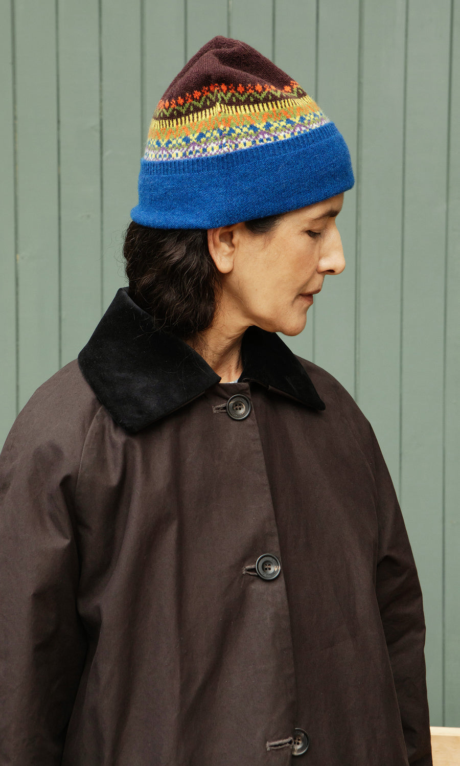 Plümo  Person in a Mull Fairisle Beanie and dark coat stands sideways against a green wooden wall.
