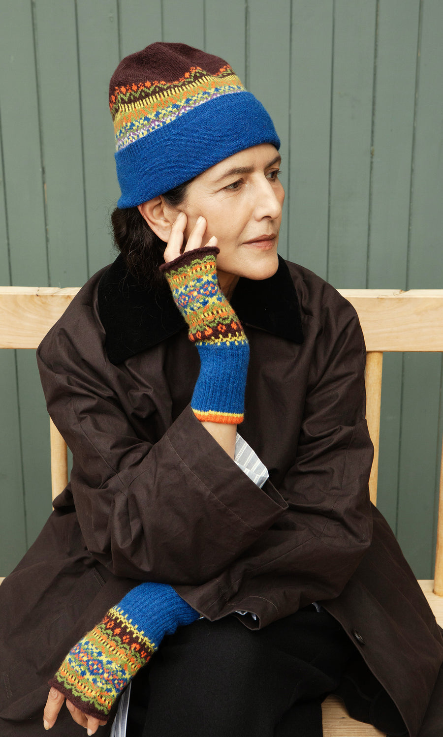 Plümo  Woman in Mull Fingerless Gloves and a fair isle knit hat sits on a bench, gazing sideways, lost in thought.
