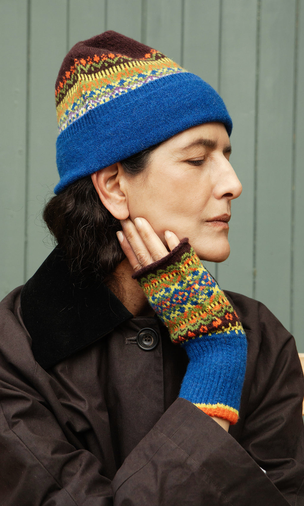 Plümo  Person in Mull Fingerless Gloves and fair isle knit hat, eyes closed, dark coat, standing by a green wall.