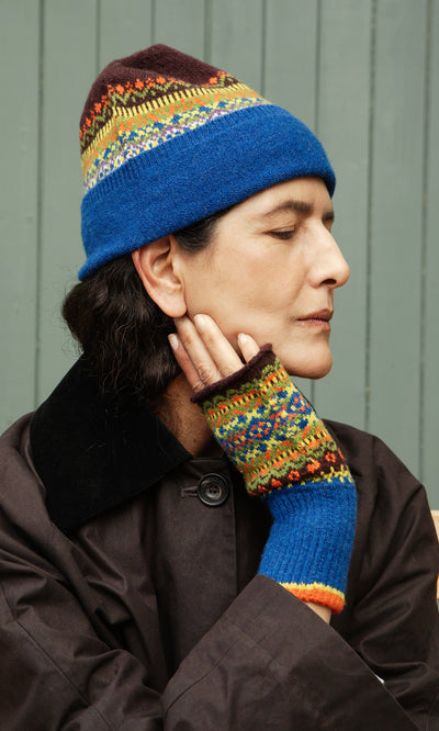 Plümo  Person wearing the Mull Fairisle Beanie, eyes closed, touching neck, in a dark coat against a green wall.
