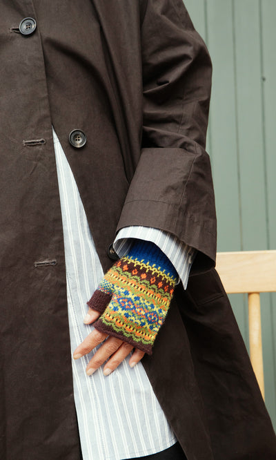 Plümo  A person in a dark coat wears Mull Fingerless Gloves, resting their hand on a wooden chair.