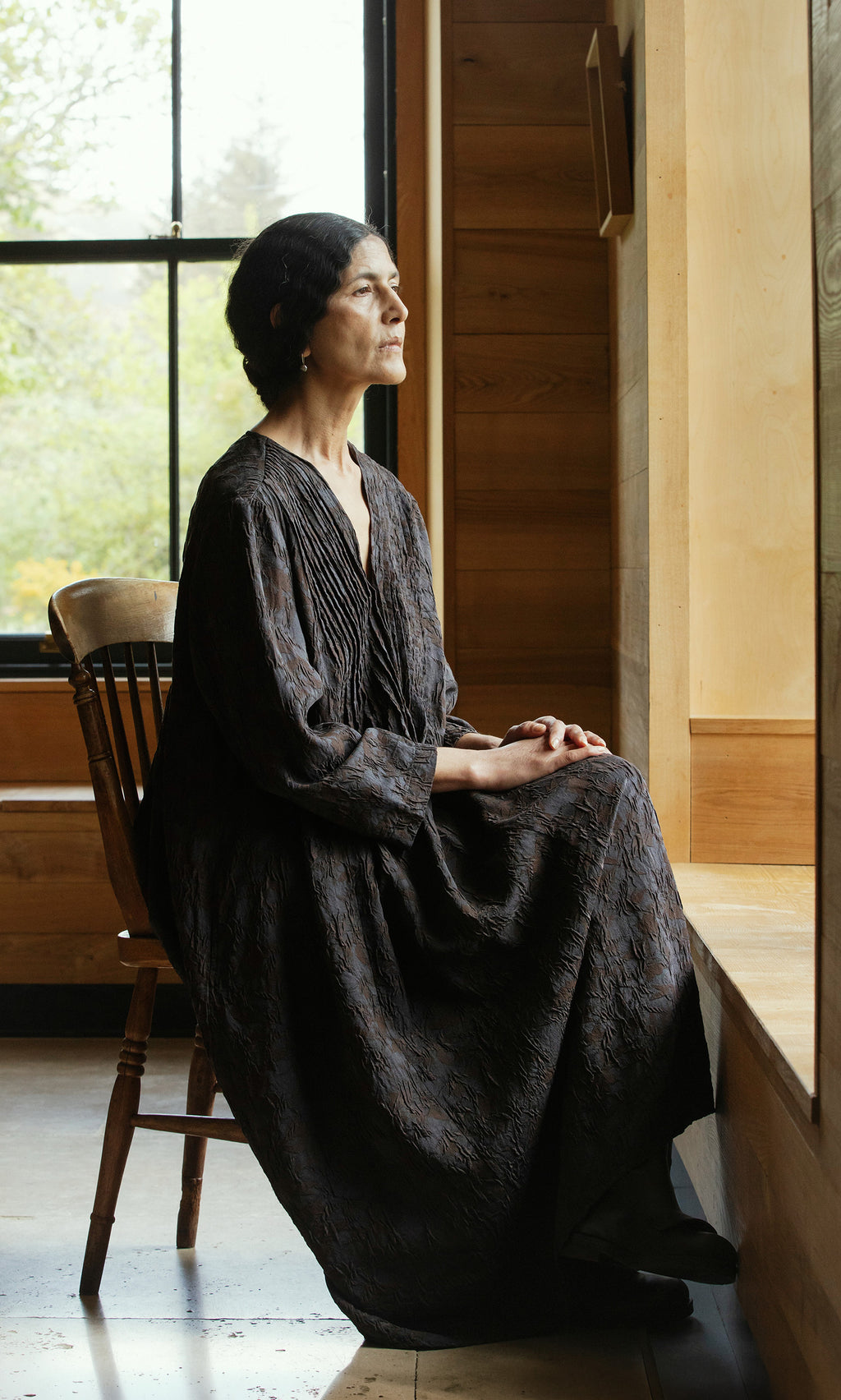 Plümo  Woman in a Mullion Dress sits on a wooden chair by a window, gazing thoughtfully outside.