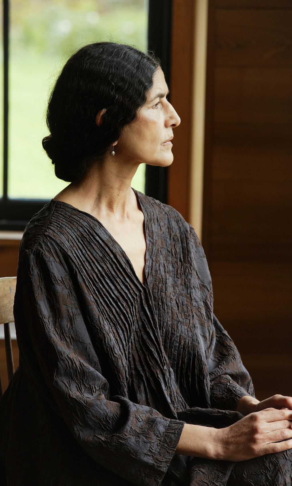 Plümo  Woman with dark hair in a bun sits in profile by a window, wearing the Mullion Dress.