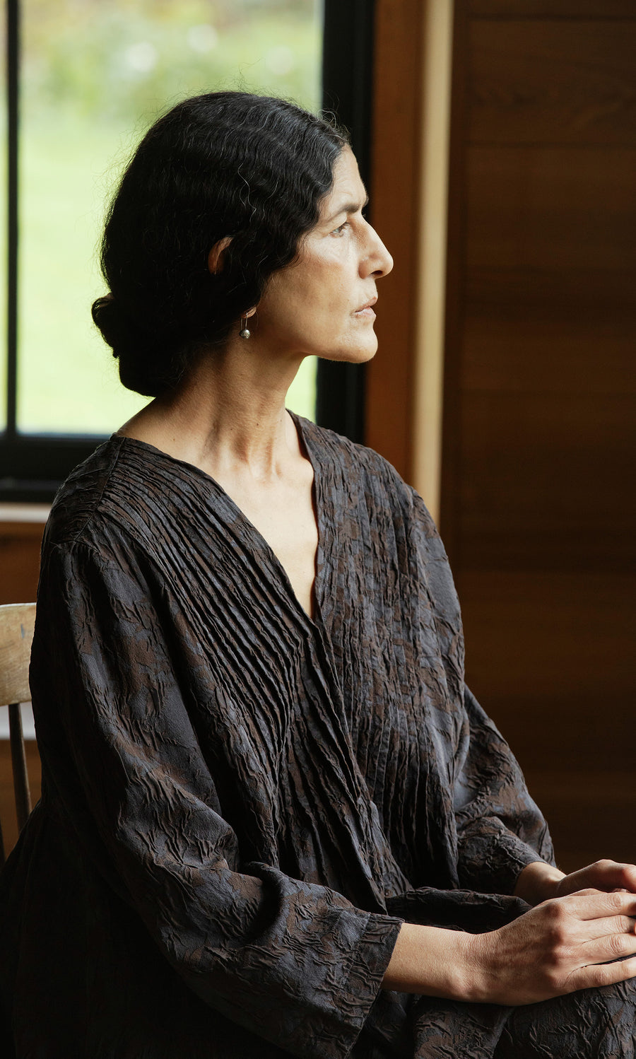 Plümo  Woman with dark hair in a bun sits in profile by a window, wearing the Mullion Dress.