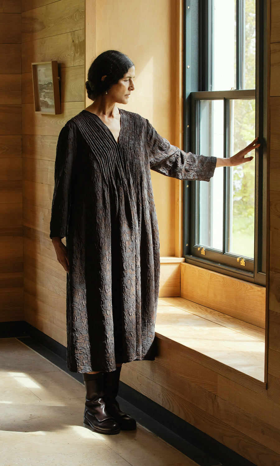 Plümo  Woman in a black Mullion Dress stands by a window, gazing out with one hand resting on the window frame.