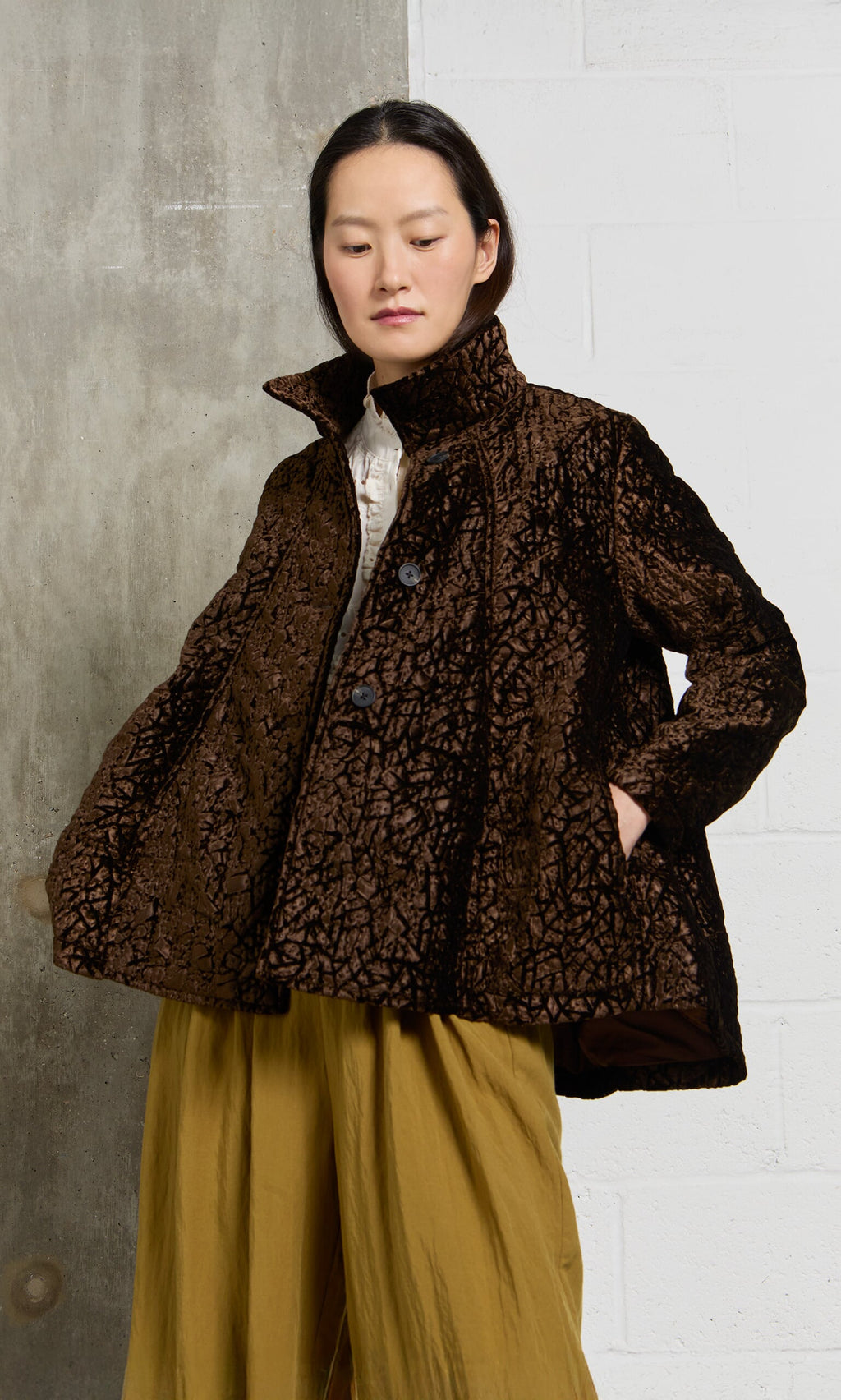 Plümo- A person in a Murano Jacket and mustard yellow pants stands against a gray and white wall.