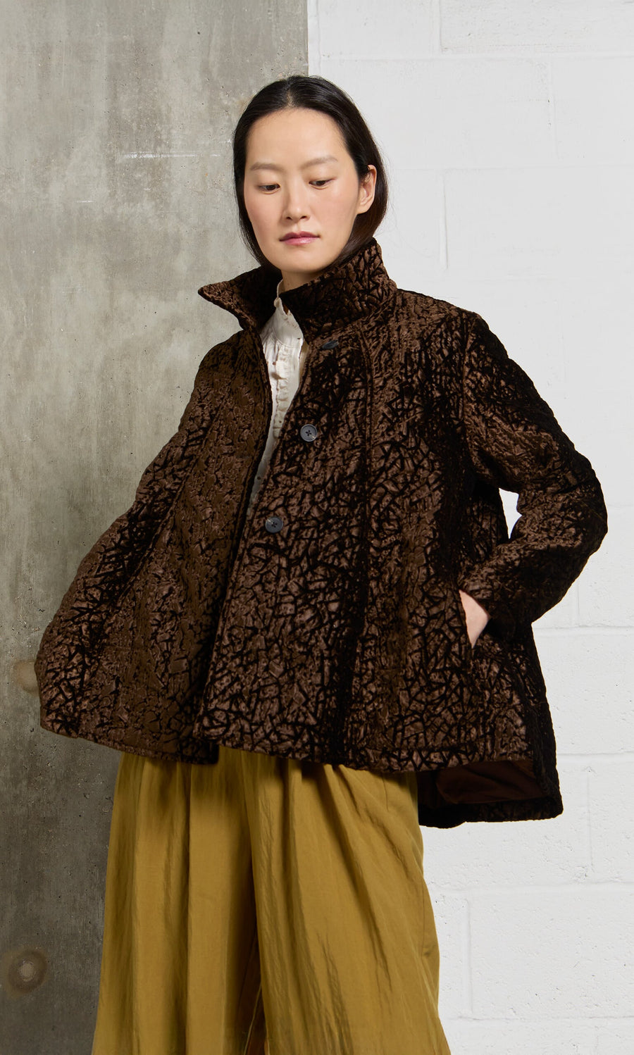 Plümo- A person in a Murano Jacket and mustard yellow pants stands against a gray and white wall.