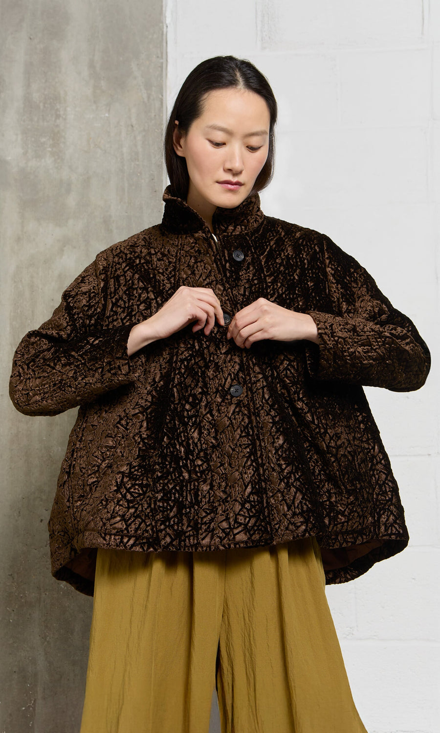 Plümo- A woman wears the Murano Jacket over a mustard-yellow dress, standing by a concrete and white wall.