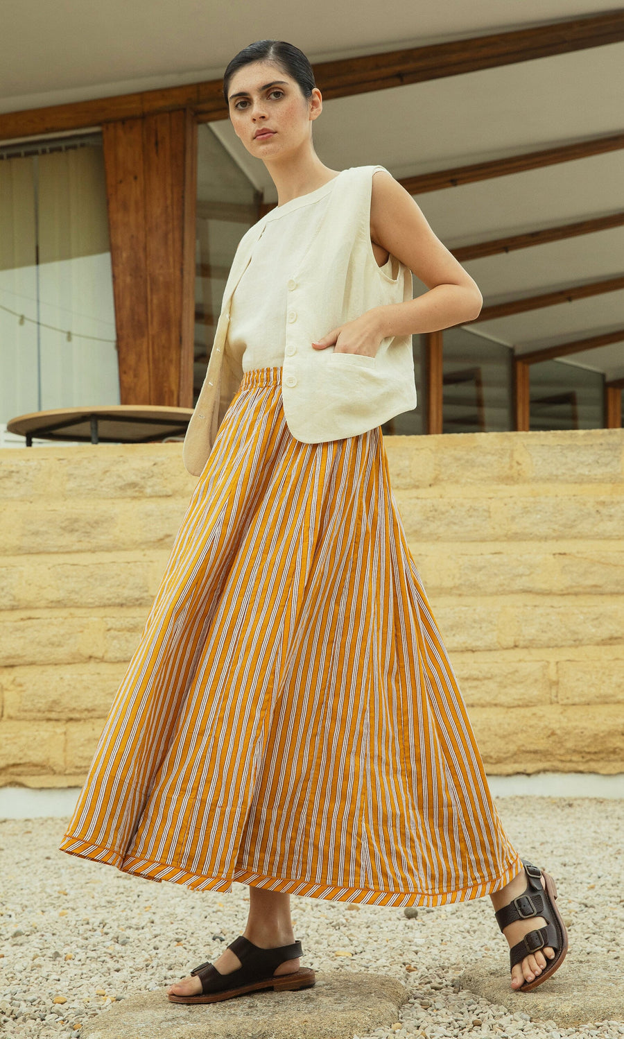 Plümo- Woman in a sleeveless beige top, Mustard Skirt with elastic waistband, and sandals stands outside a modern building.